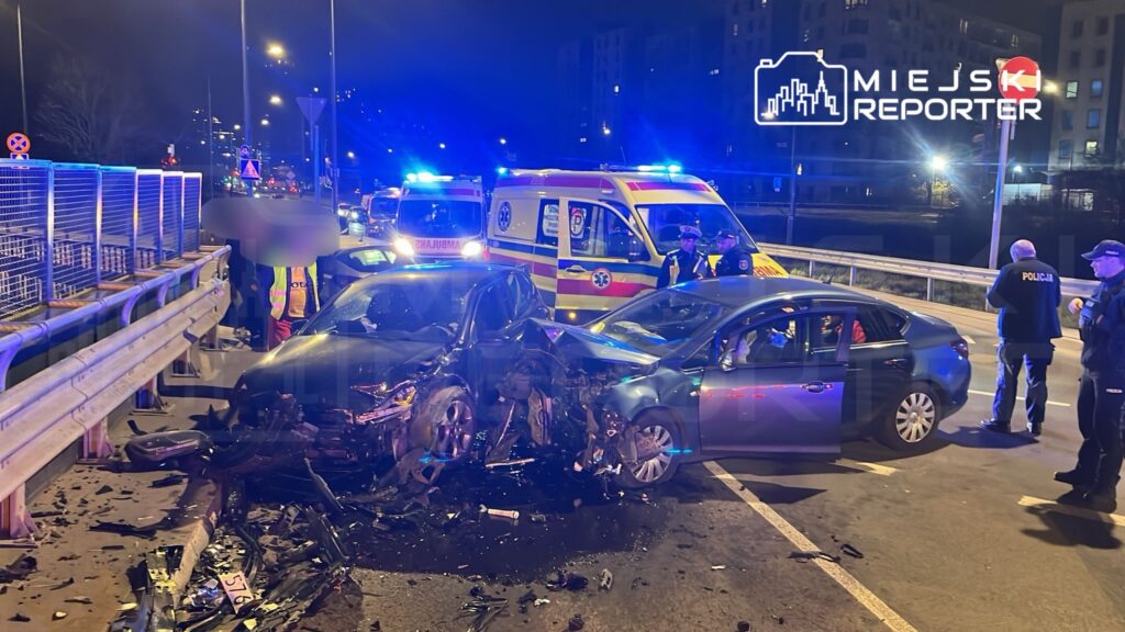 W nocy na drodze doszło do wypadku, w którym zderzyły się dwa samochody. W tle widać karetki pogotowia i policjantów zabezpieczających miejsce zdarzenia.