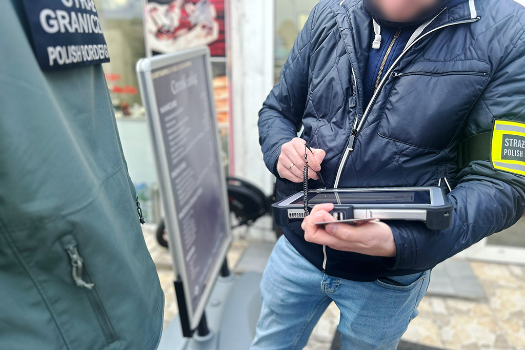 Mężczyzna w niebieskiej kurtce trzyma tablet i notuje informacje obok znaku informacyjnym w mieście.