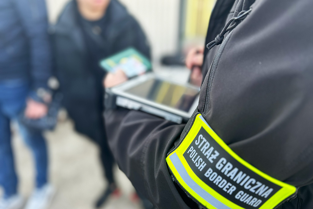 Funkcjonariusz Straży Granicznej z tabletem w ręku, kontrolujący dokumenty w otoczeniu innych osób.