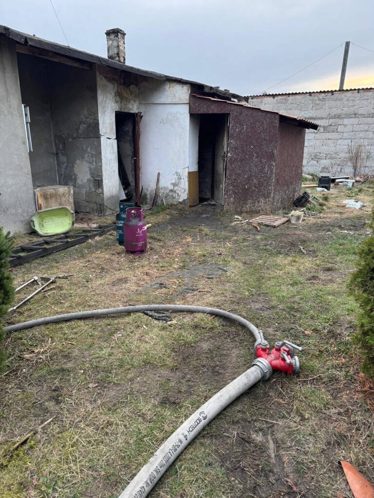 Zniszczony budynek z odrapaną elewacją, obok wąż strażacki i butla gazowa na trawie.