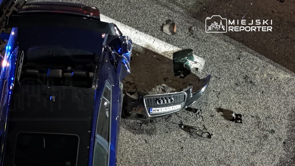 Zniszczony samochód osobowy leży na drodze, obok fragmenty karoserii i szkła, w nocy.