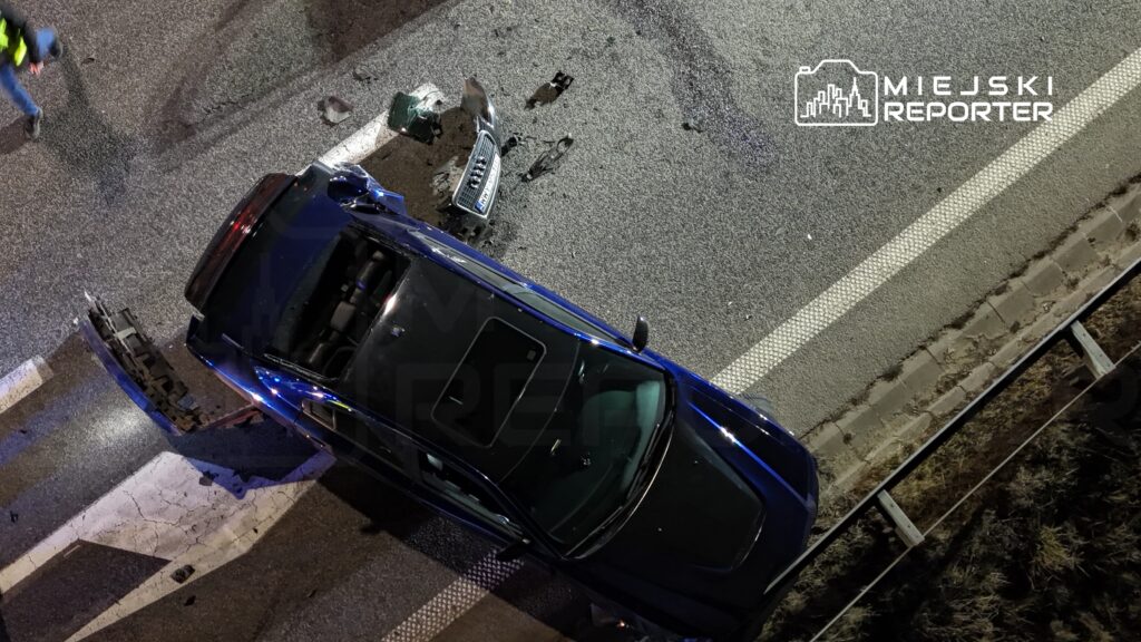 Czarny samochód leży na boku na drodze, wokół rozrzucone fragmenty pojazdu, a obok stoi osoba w odblaskowej kamizelce.