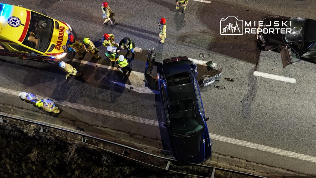 Grupa ratowników oraz policjantów pracuje na miejscu wypadku drogowego, obok uszkodzonego samochodu na jezdni.