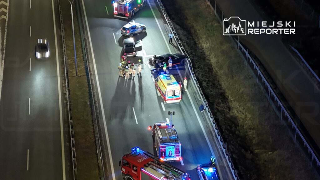 Grupa ratowników medycznych i strażaków udziela pomocy poszkodowanym na drodze po wypadku samochodowym.