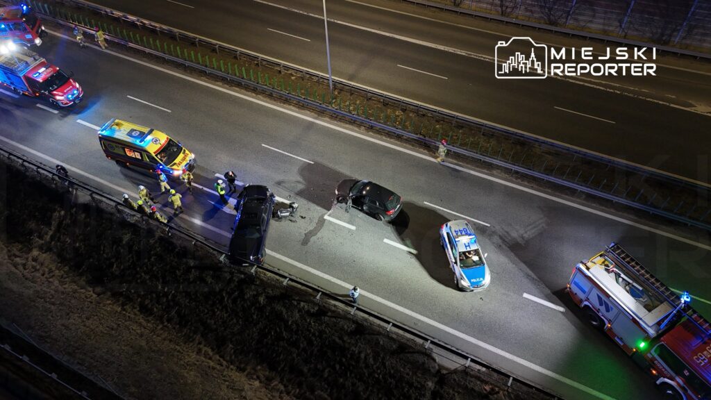 Na drodze leży uszkodzony czarny samochód, obok niego pracują strażacy i policja, a w tle widać pojazdy ratunkowe.
