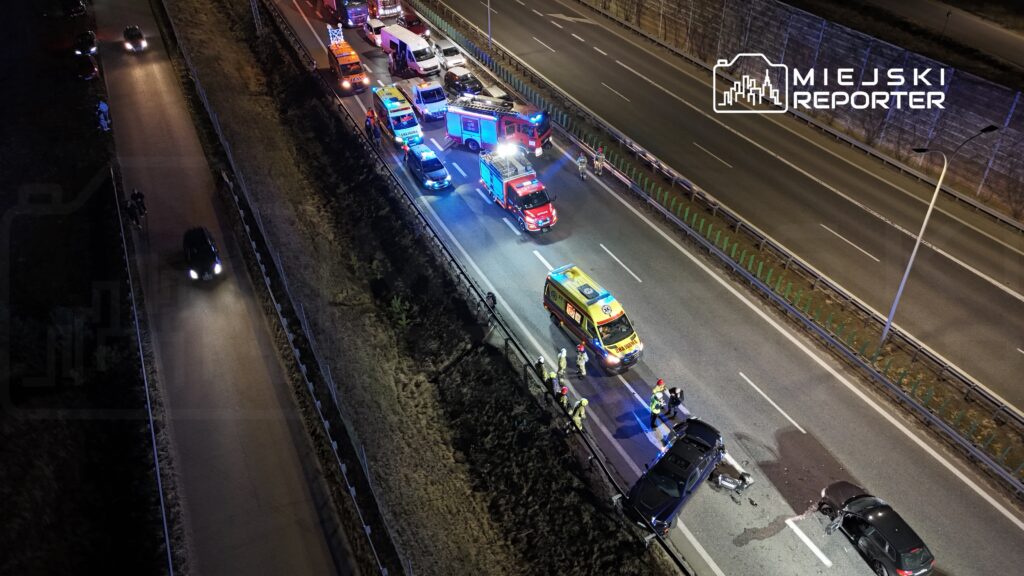 Na drodze autostradowej zderzenie dwóch samochodów; w pobliżu pracują służby ratunkowe i strażacy.