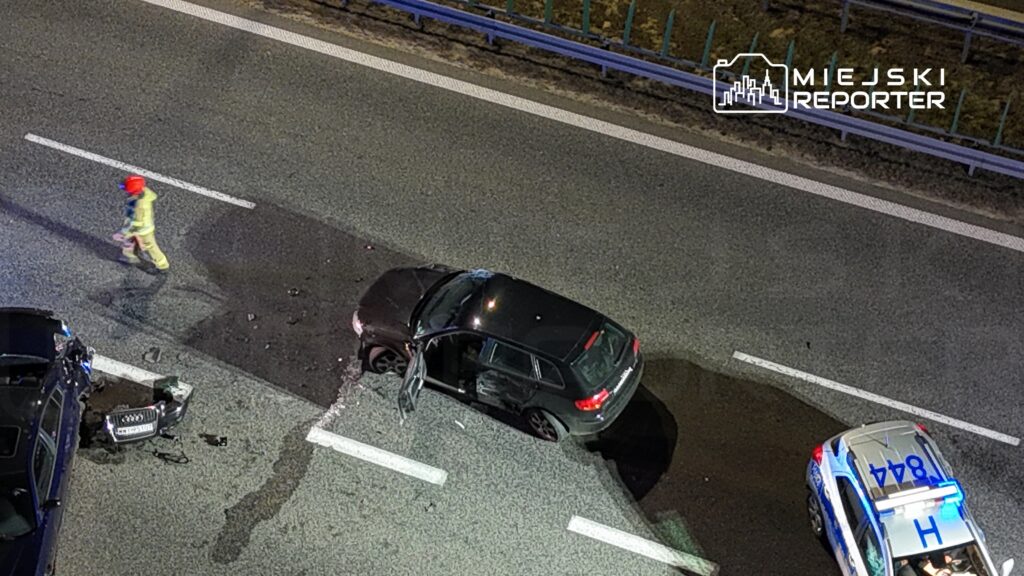 Strażak w żółtym uniformie idzie wzdłuż drogi obok uszkodzonego czarnego samochodu, a w tle widoczny radiowóz.