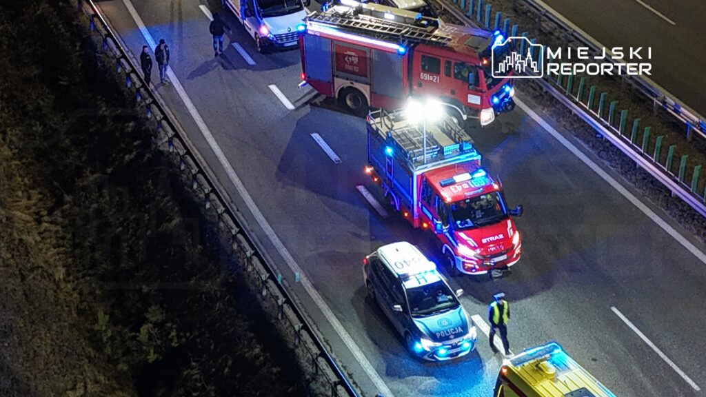 Grupa ludzi obserwująca działania służb ratunkowych na autostradzie, gdzie zjechały pojazdy straży pożarnej i policji.