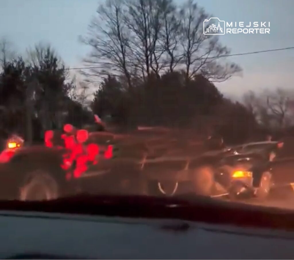 Pojazd z drewnem zatrzymany na drodze o zmierzchu, w tle drzewa i słabe oświetlenie.