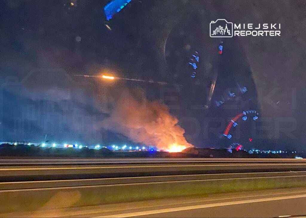 Ogień i dym unoszą się nad terenem w nocy, w tle widoczne są światła pojazdów na drodze.