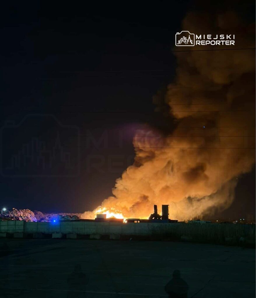 Ognisko pożaru na wysypisku, z unoszącym się gęstym dymem w nocy, oświetlonym przez niebieskie światła straży pożarnej.