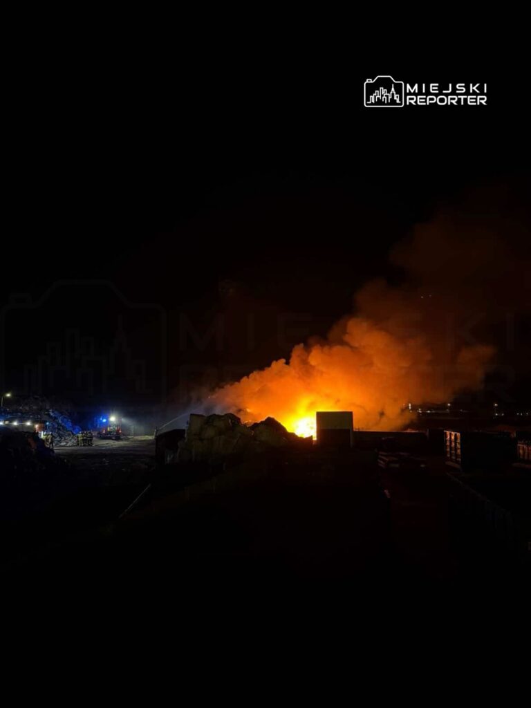 Ognisko pożaru wydobywające się z sterty odpadów nocą, z widocznymi w tle wozami strażackimi.