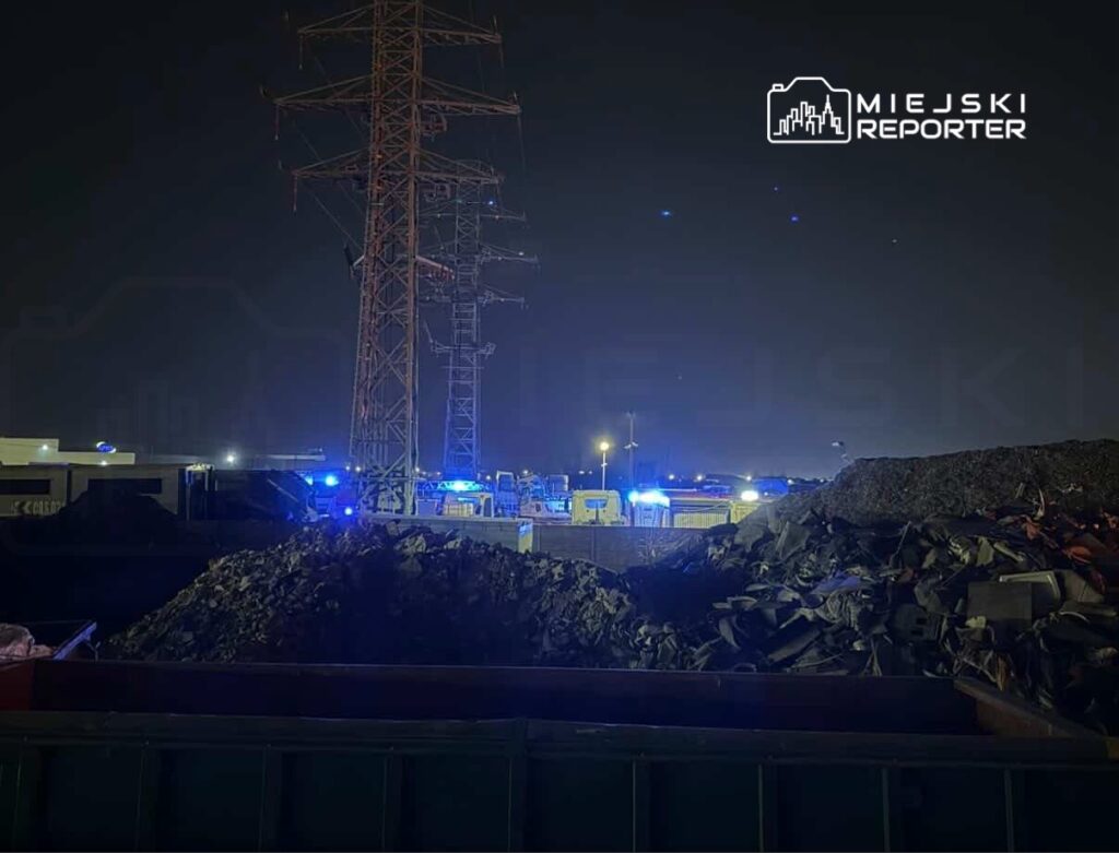 Na placu budowy w nocy, w tle widać oświetlone pojazdy służb ratunkowych oraz wieżę energetyczną.