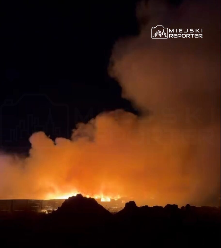 Ogromne płomienie i gęsty, pomarańczowy dym unoszą się w nocy nad terenem przemysłowym.