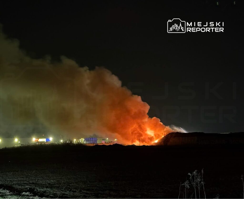 Ogromny pożar wytwarzający gęsty dym, widoczny w nocy na tle oświetlonych budynków.