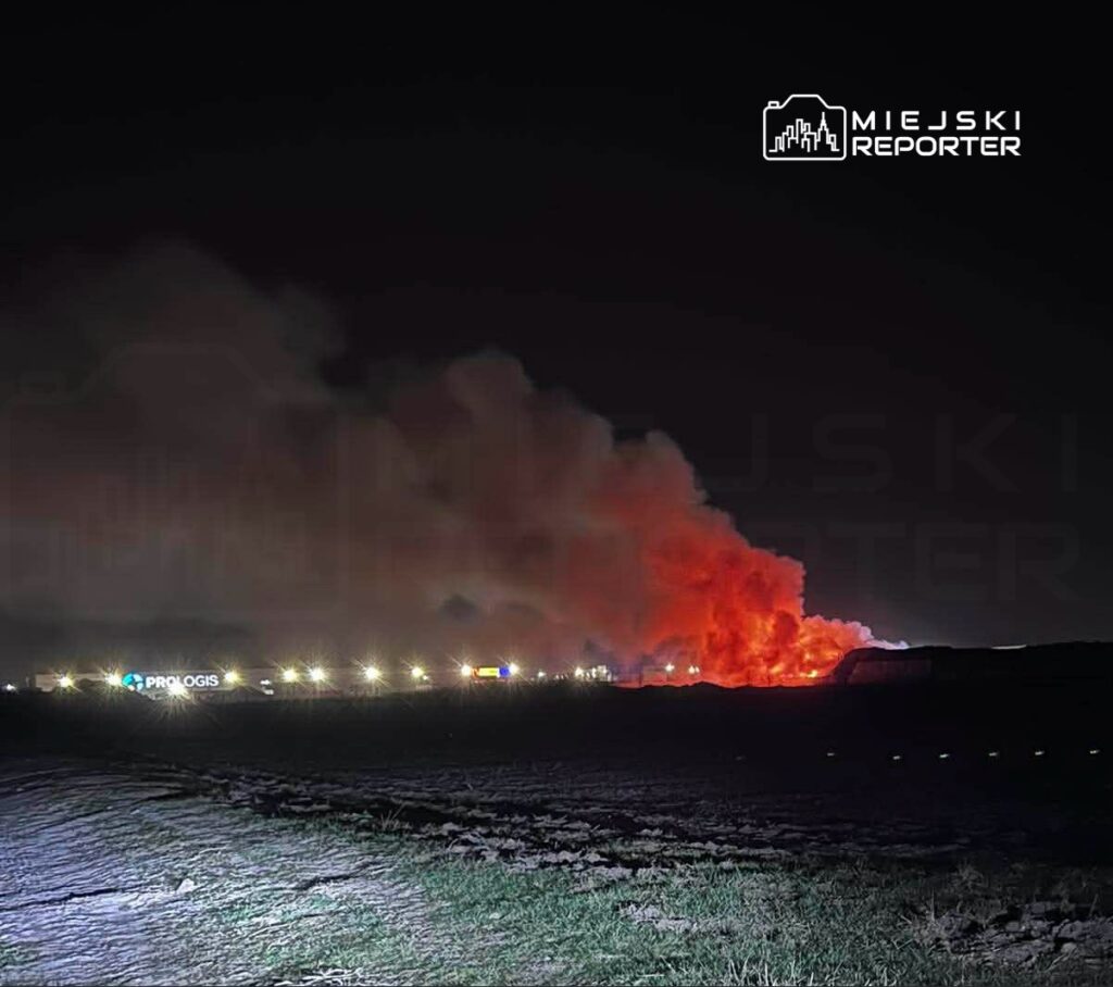 W nocy widać intensywny pożar, z którego unosi się gęsty dym i płomienie, w tle widoczne oświetlenie przemysłowe.