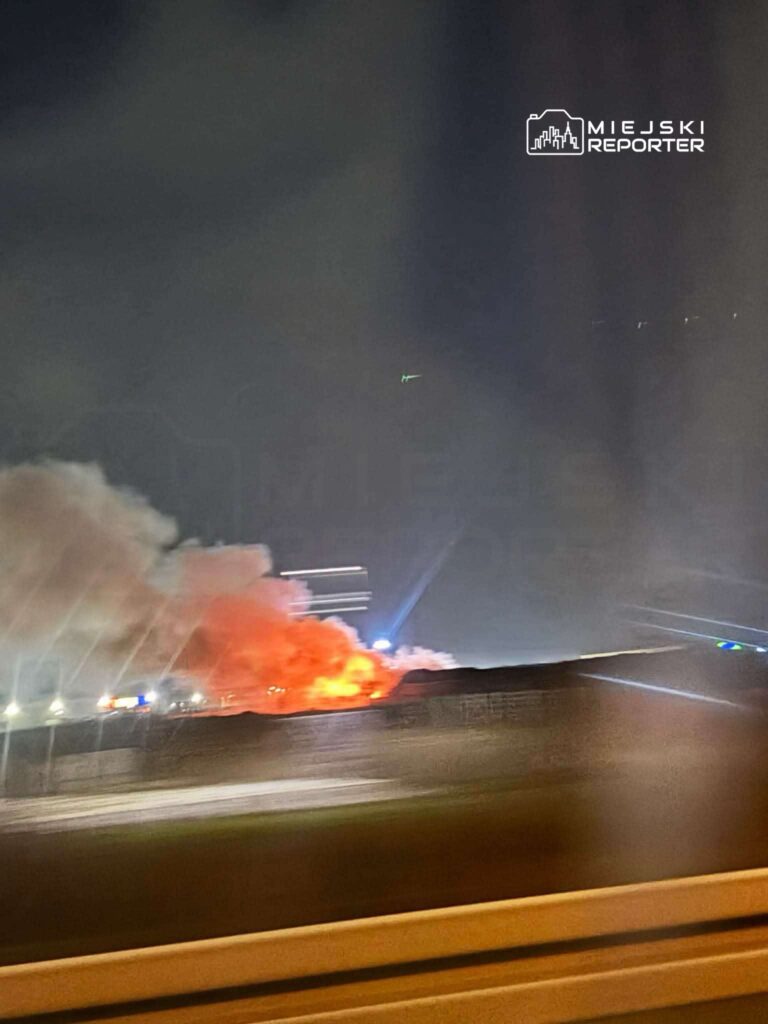 Intensywne płomienie i gęsty dym unoszą się nad terenem przemysłowym w nocy, w tle widoczne światła służb ratunkowych.