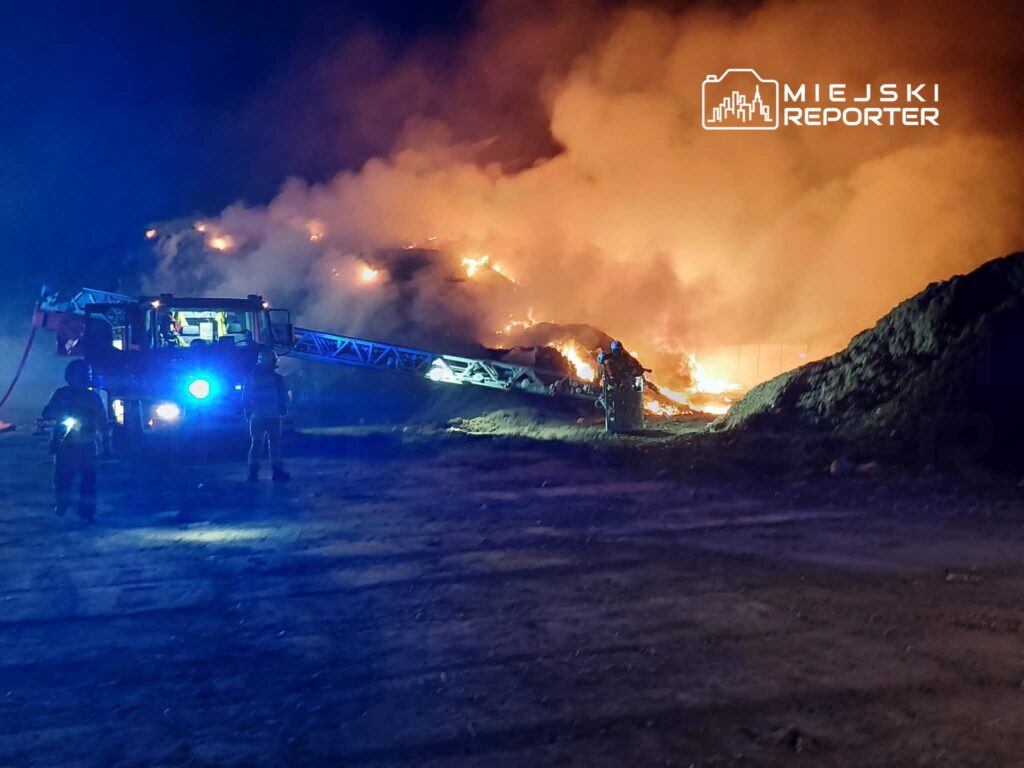 Strażacy gaszą pożar na wysypisku, używając wozu strażackiego z drabiną i wężem, w otoczeniu dymu i płomieni.