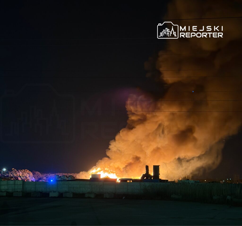 W nocy widoczny jest pożar na wysypisku, z którego wydobywa się gęsty dym i płomienie, otoczone ciemnym niebem.