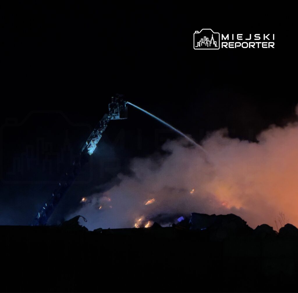 Strażak na wysięgniku gasi pożar w otoczonym dymem terenie, w nocy.