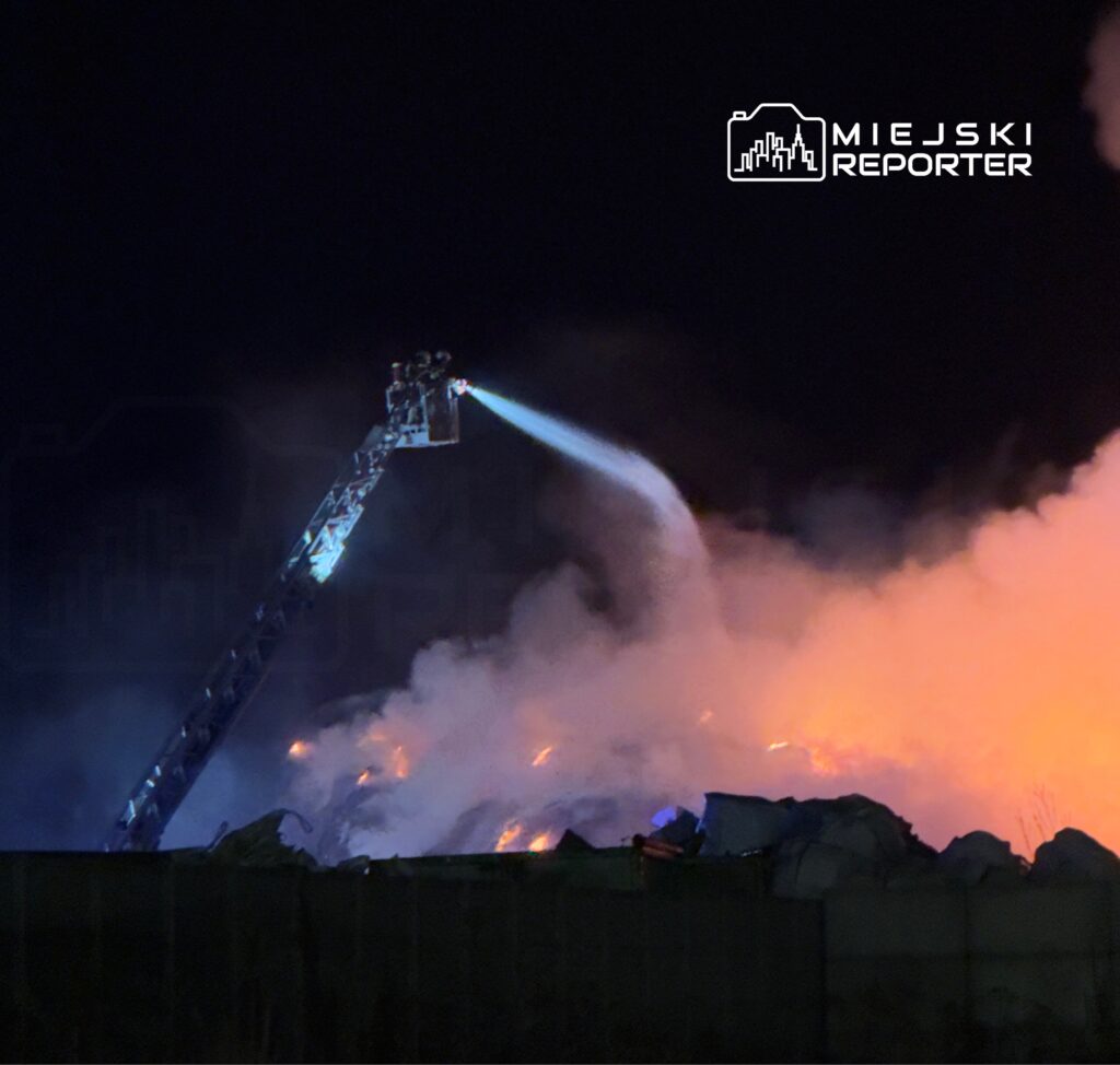 Strażak na wysięgniku gaśniczym polewa wodą płonące zgliszcza w nocy, otoczone gęstym dymem.