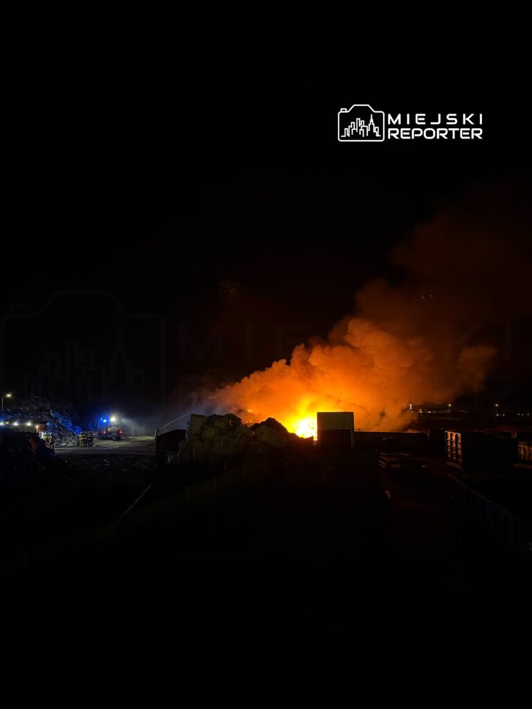 Ognisko pożaru otoczone dymem w nocy, z widocznymi w tle wozami strażackimi i stosami materiałów.