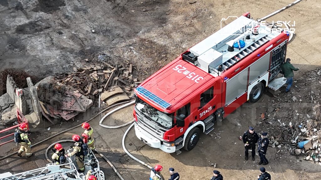 Czerwony wóz strażacki stoi na placu budowy, a w pobliżu strażacy w hełmach rozmawiają z policjantami.