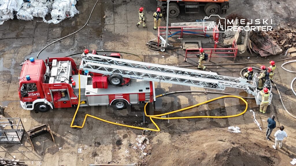Strażacy w hełmach i mundurach przygotowują sprzęt na terenie zakładu, obok wozu strażackiego z wysięgnikiem.