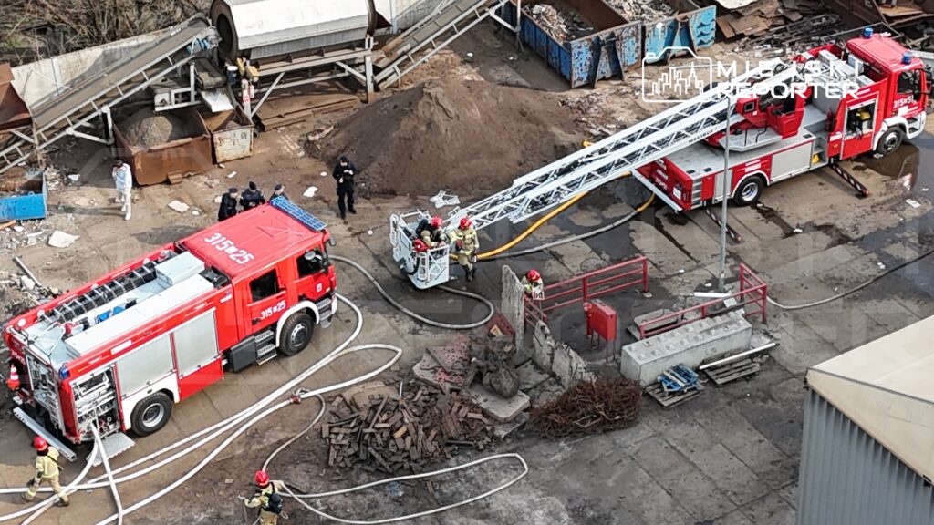Strażacy w hełmach obsługują drabinę strażacką obok wozu strażackiego na terenie zakładu przemysłowego.