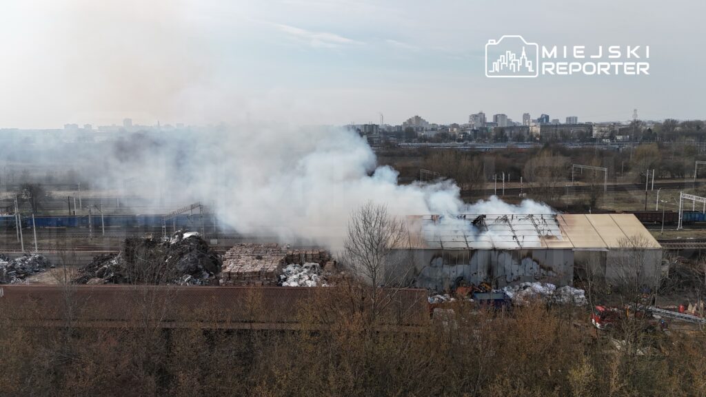 Dym unoszący się z płonącego budynku magazynowego w przemysłowej okolicy, w tle widoczne linie kolejowe i miasto.