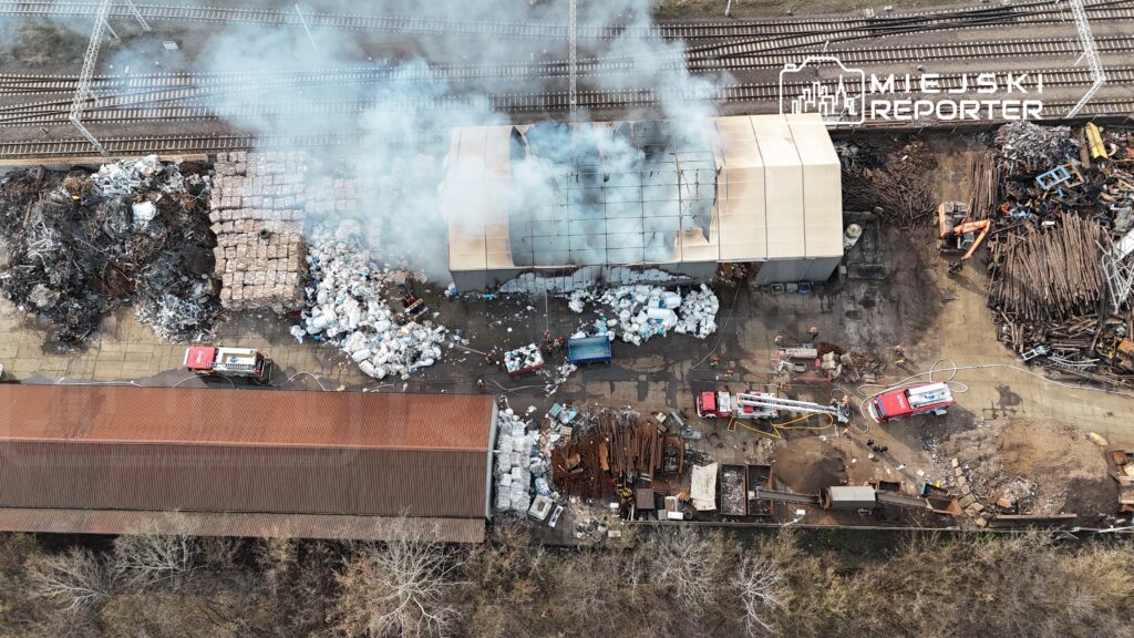 Dym unoszący się nad halą przemysłową, w której strażacy gaszą pożar, otoczoną stertami odpadów metalowych i plastikowych.