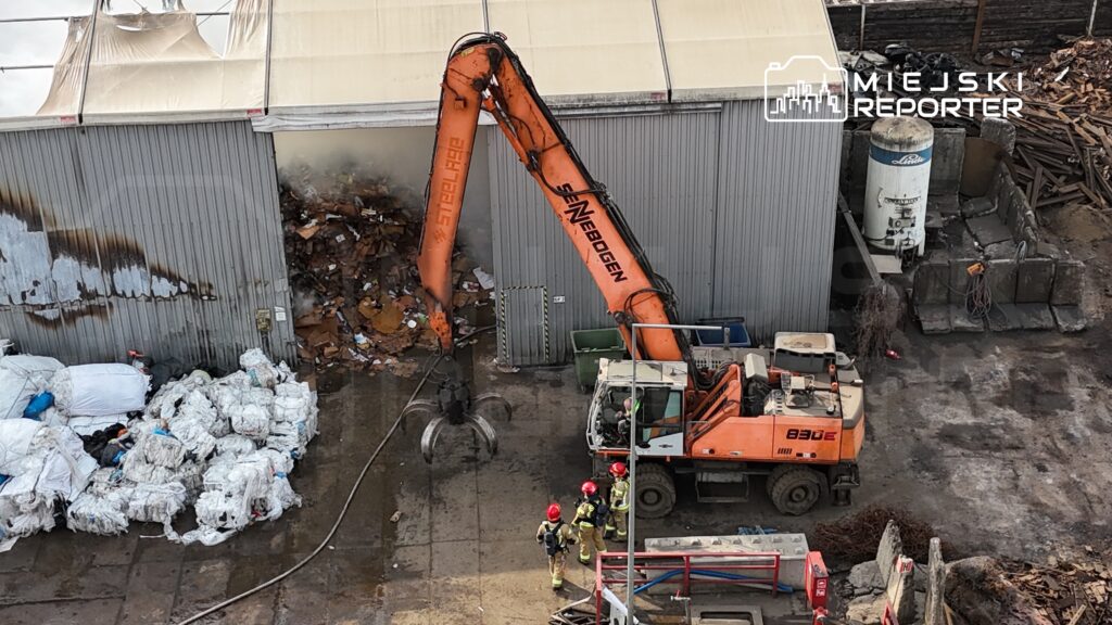 Wielka maszyna hydrauliczna podnosi odpady w pobliżu hali przemysłowej, a obok stoi zespół strażaków w hełmach.