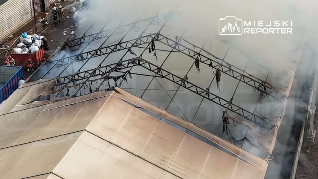 Strażacy w hełmach gaszą pożar w dymiącym budynku z uszkodzoną konstrukcją dachową.