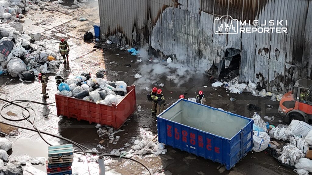 Grupa strażaków w hełmach, kontrolująca dym unoszący się z wysypiska śmieci obok kolorowych kontenerów.