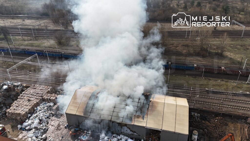 Gęsty dym unoszący się z dachu magazynu na tle torów kolejowych oraz składowiska odpadów.
