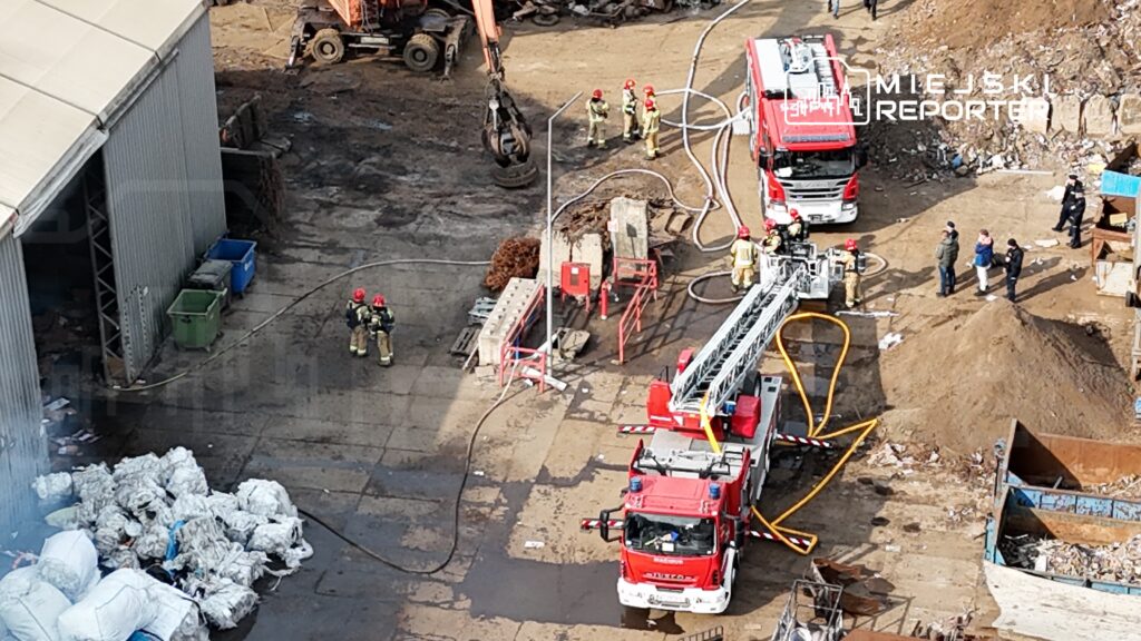 Strażacy w hełmach i mundurach rozstawiają sprzęt gaśniczy na terenie zakładu przetwarzania odpadów.