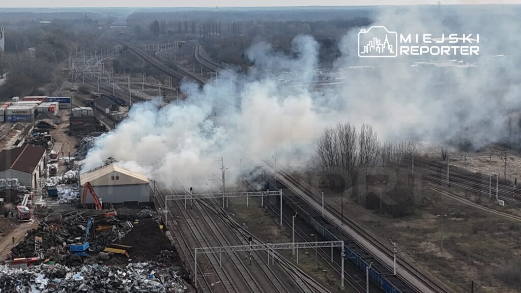 Gęsty dym unoszący się nad halą w pobliżu torów kolejowych, obok strefy złomowej.