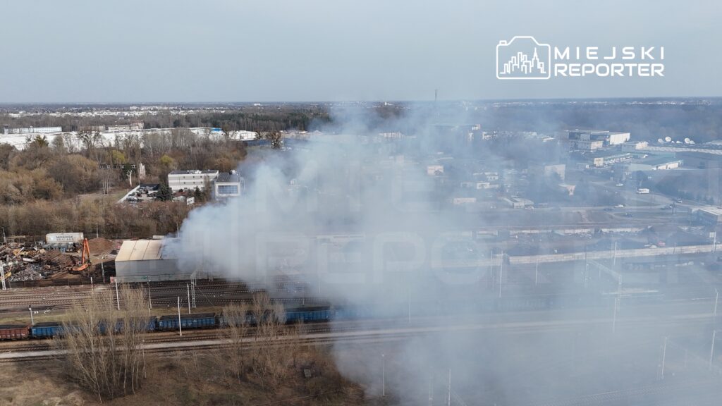 Dym unoszący się nad torami kolejowymi w przemysłowej okolicy, w tle widoczne budynki i drzewa.