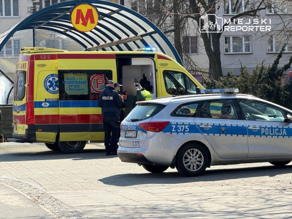 Grupa policjantów rozmawia z ratownikami medycznymi przy karetce na przystanku metra w mieście.