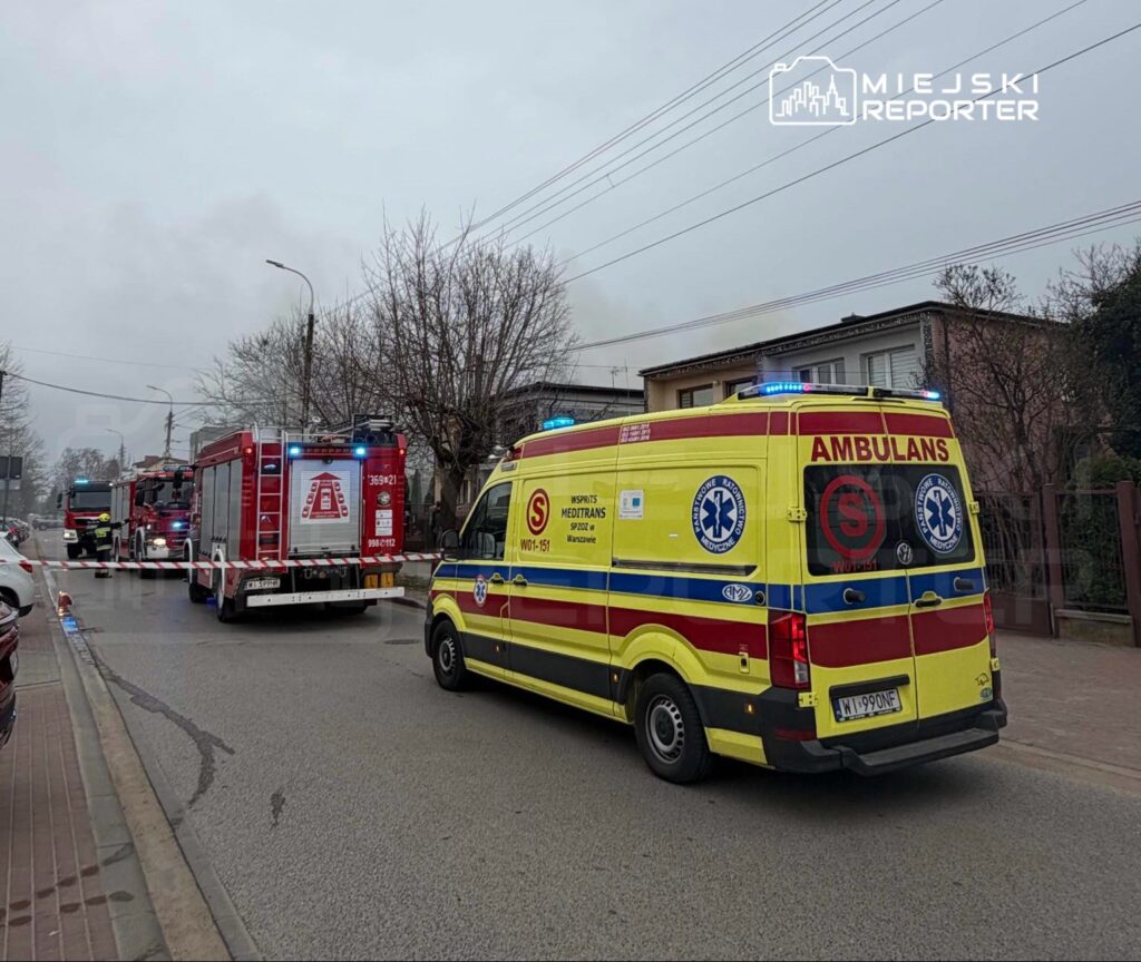 Na ulicy stoją karetka pogotowia oraz wozu strażackiego, w tle widoczny dym oraz zablokowany ruch.