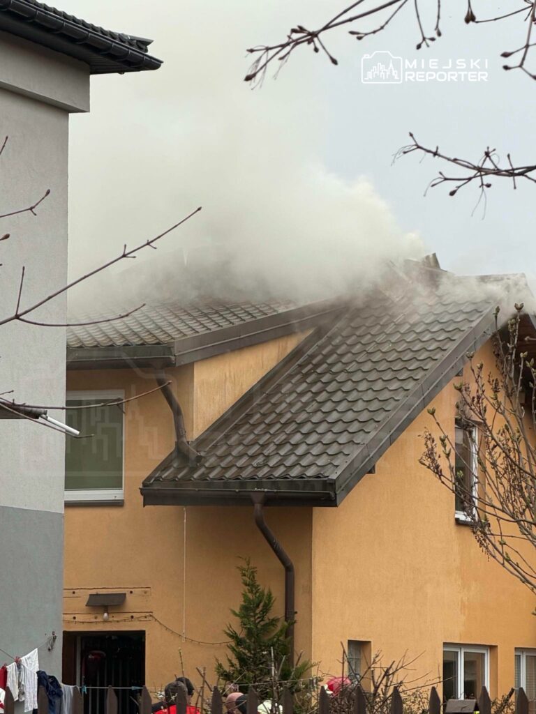 Dym wydobywający się z dachu budynku mieszkalnego w otoczeniu drzew i suszących się ubrań na lince.