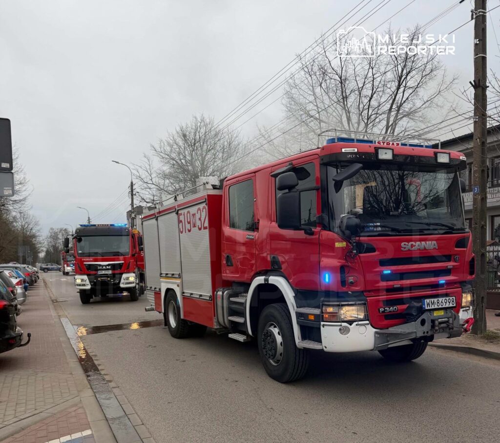 Dwa wozy strażackie w czerwonym kolorze poruszają się ulicą, obok zaparkowanych samochodów.