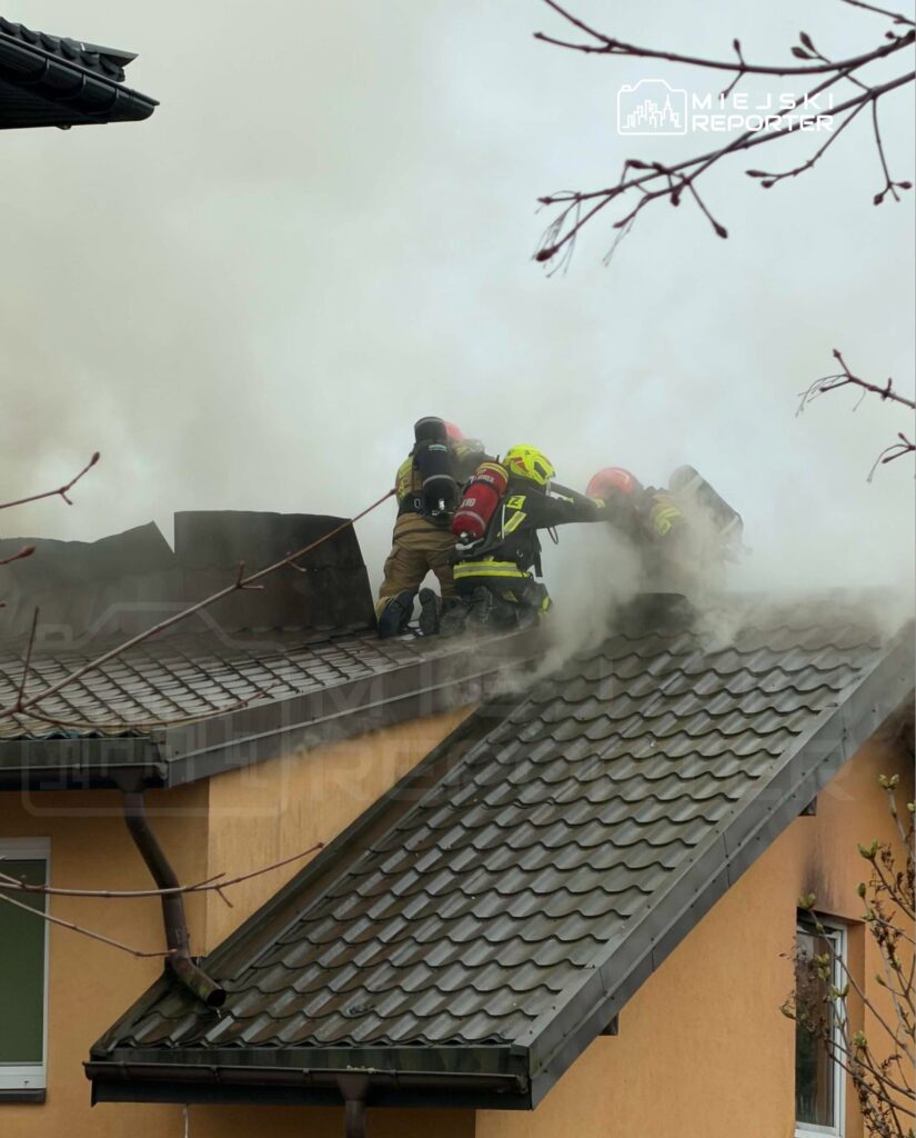 Czterech strażaków w sprzęcie ochronnym walczy z pożarem na dachu budynku, otoczonym gęstym dymem.