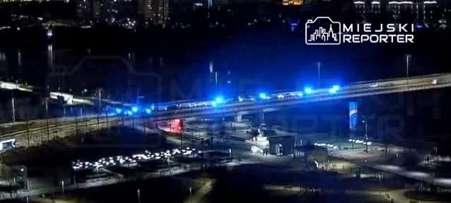 Nocna scena z mostem oświetlonym niebieskimi światłami, w tle widoczne budynki i parking.