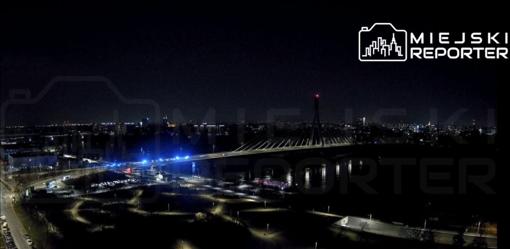 Most z oświetleniem w niebieskich barwach, łączący dwa brzegi rzeki, otoczony miastem nocą.