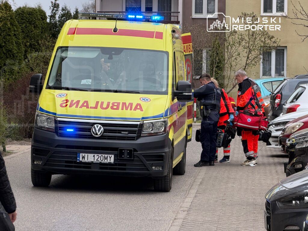 Zespół ratowników medycznych w czerwonych strojach podchodzi do ambulansu zaparkowanego na osiedlowej ulicy.