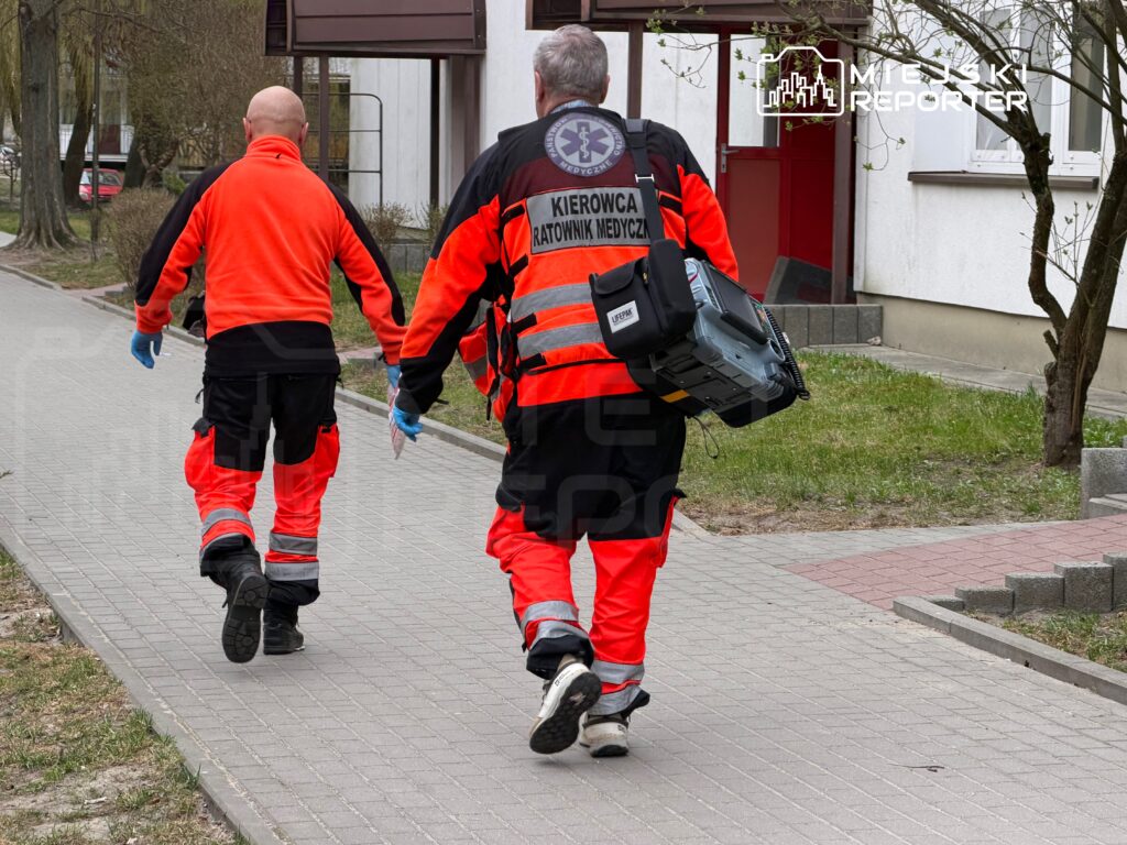 Dwaj ratownicy medyczni w pomarańczowych uniformach idą chodnikiem w kierunku budynku, niosąc sprzęt medyczny.