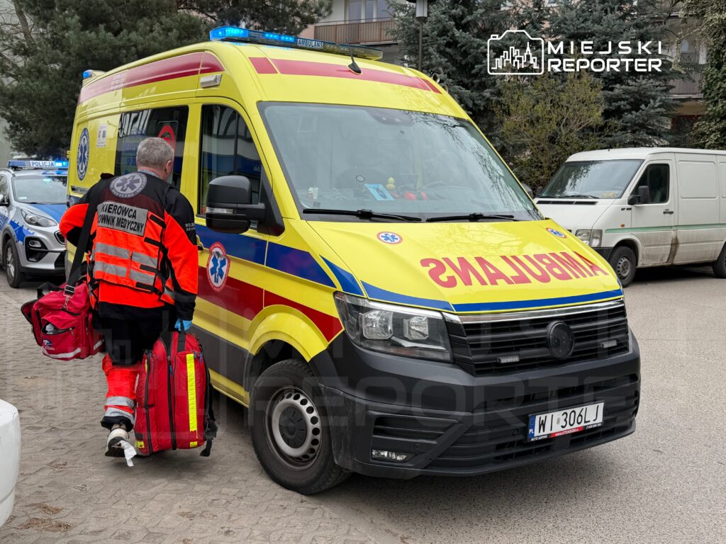 Ratownik medyczny w odblaskowym ubraniu niesie torby do karetki pogotowia zaparkowanej na ulicy.