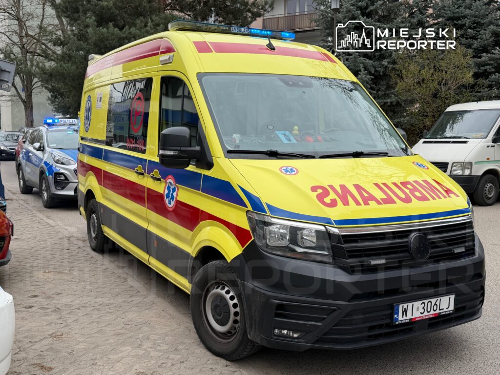 Karetka pogotowia w kolorze żółto-czerwonym zatrzymana na ulicy, obok pojazdów policyjnych z włączonymi sygnałami świetlnymi.