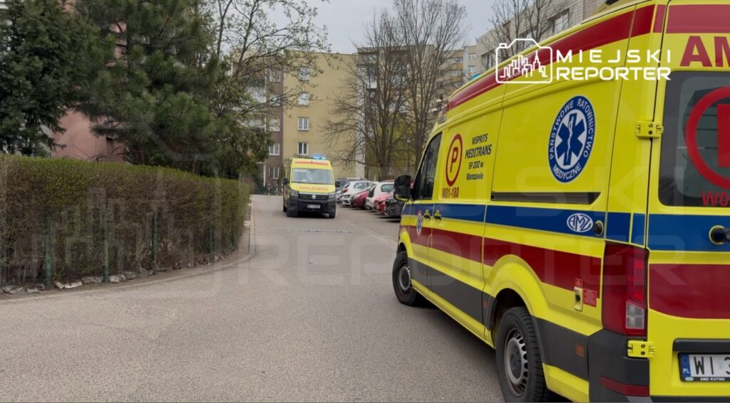 Dwa karetki pogotowia zaparkowane na wąskiej ulicy, otoczone drzewami i budynkami mieszkalnymi.